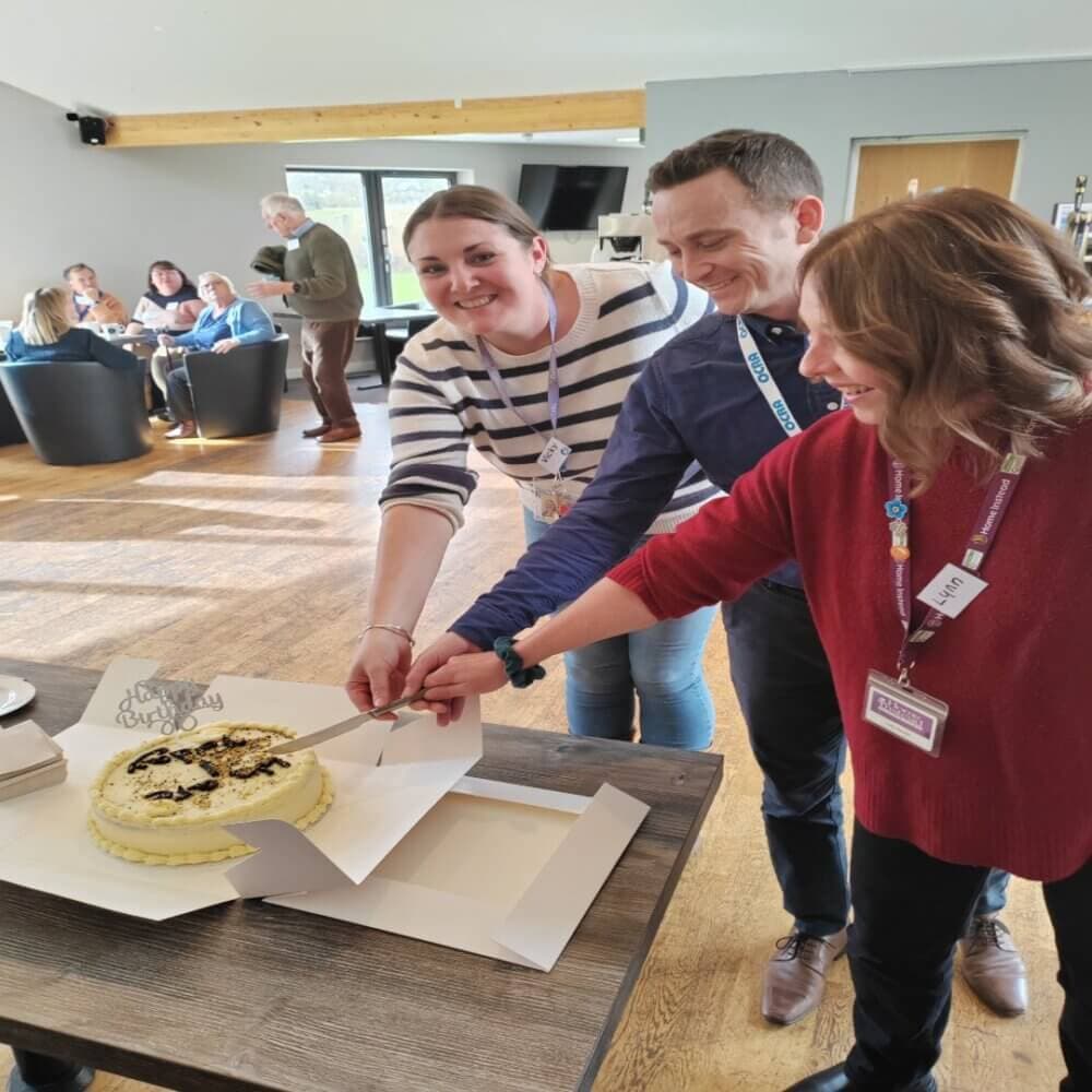 Three people smile while cutting a cake together in a room with others seated and talking in the background. - Home Instead
