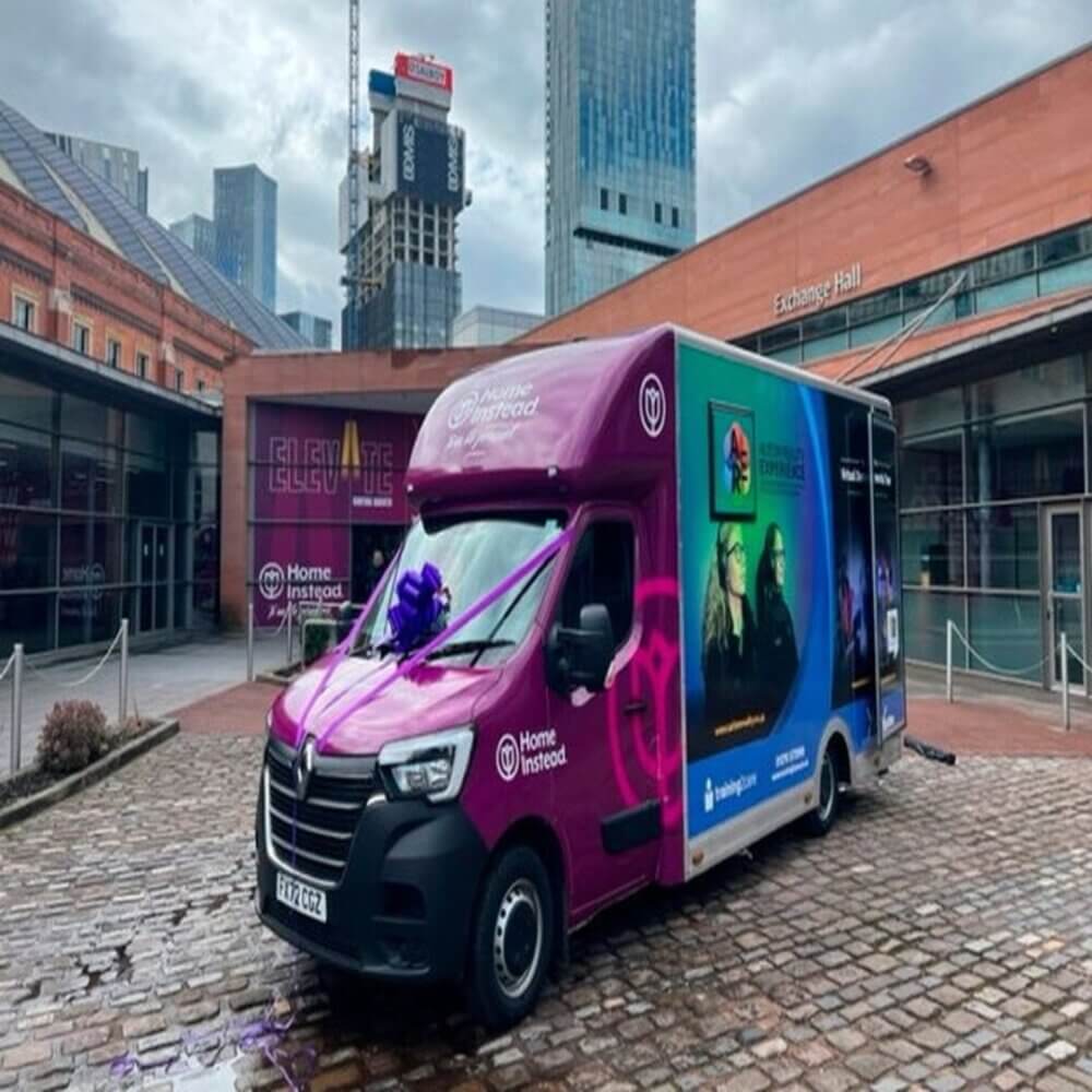 Colorful van with a purple and blue design parked in a city square near modern buildings with a cloudy sky. - Home Instead