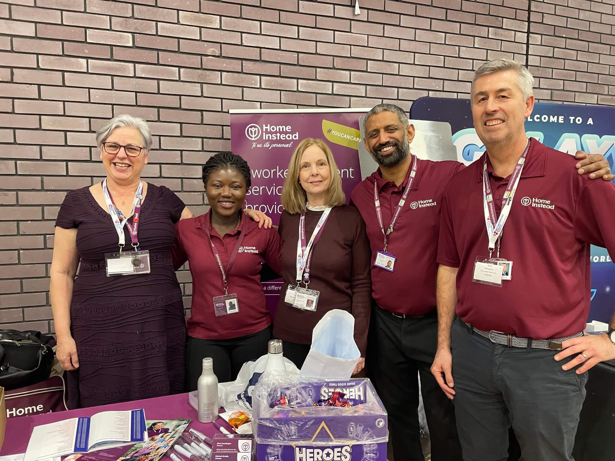 A group of five smiling people in matching maroon shirts pose at a booth with promotional materials. - Home Instead