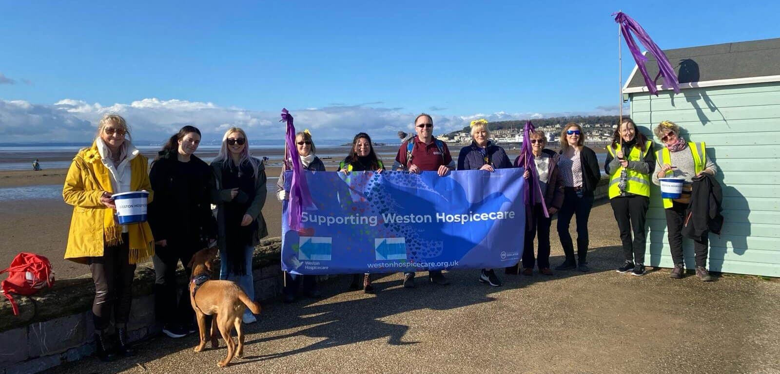 A group of people on a beach holding a "Supporting Weston Hospicecare" banner, with a dog and fundraising buckets. - Home Instead