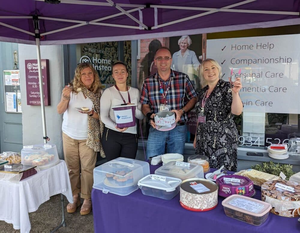Four people standing under a purple canopy with tables of baked goods for a charity bake sale. - Home Instead