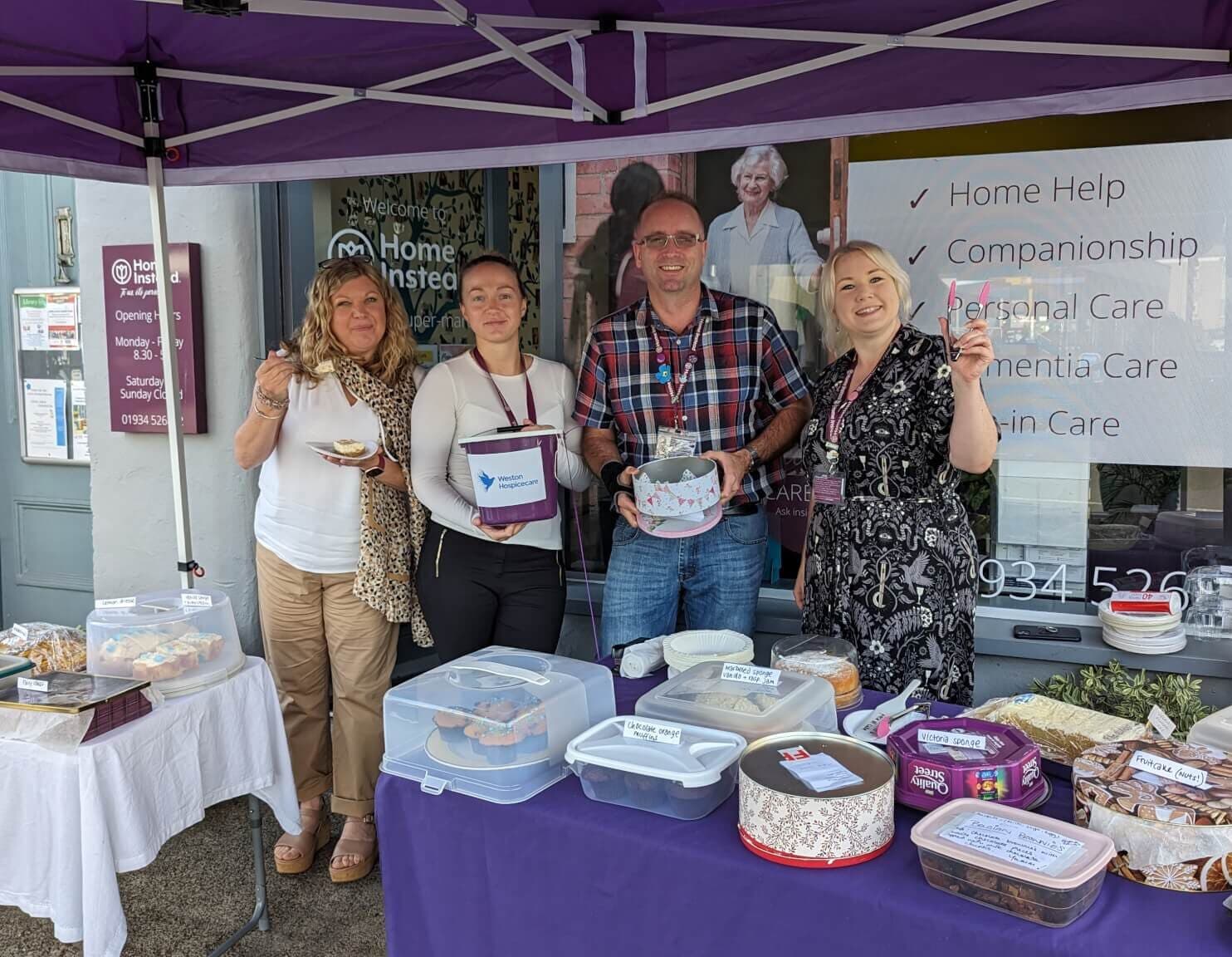 Four people standing under a purple canopy with tables of baked goods for a charity bake sale. - Home Instead