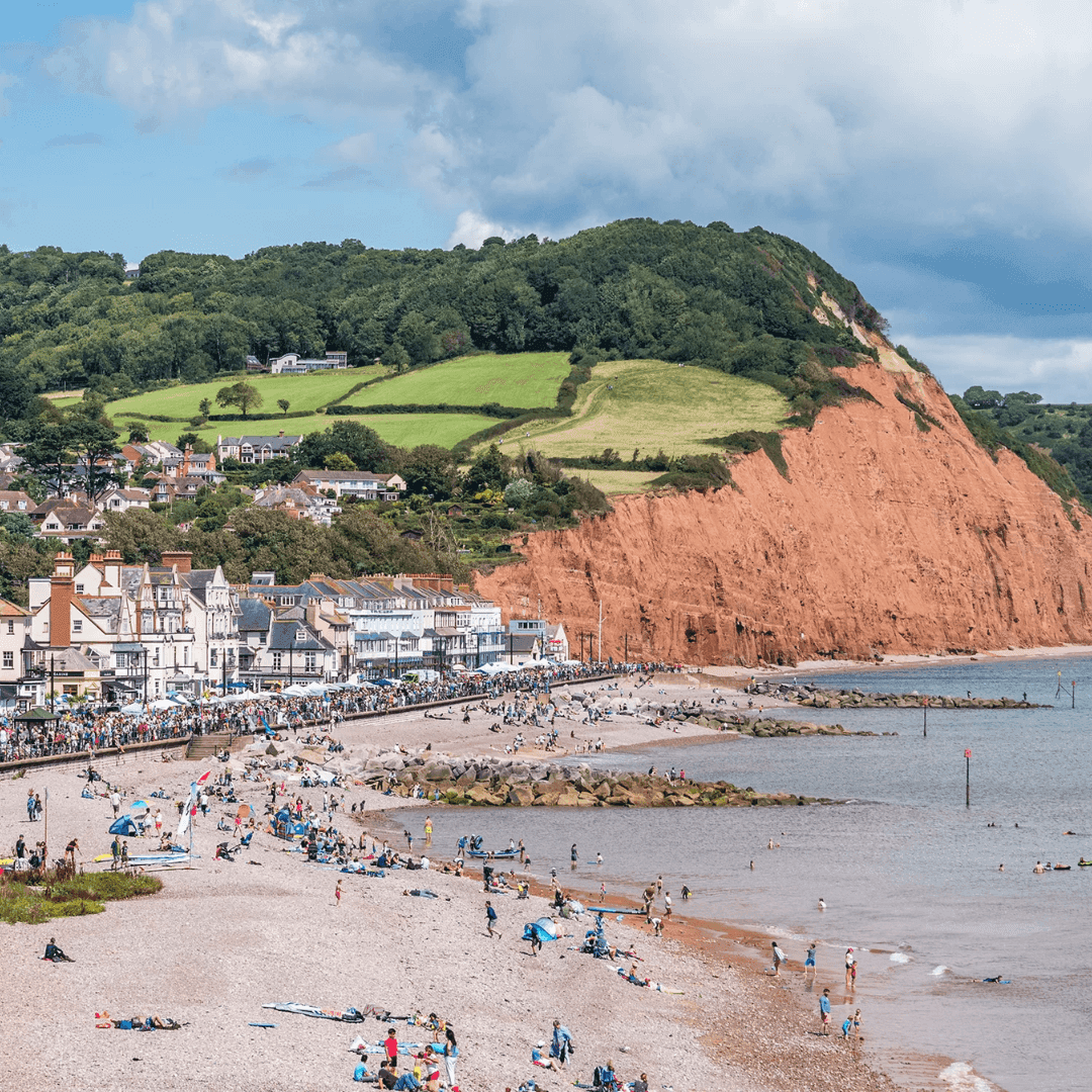 Scenic beach with visitors, coastal houses, rugged cliffs, and green hills under a partly cloudy sky. - Home Instead