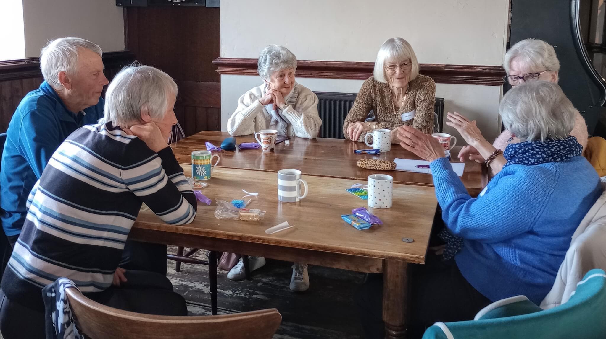 A group of elderly people sitting around a wooden table, engaged in conversation and drinking tea. - Home Instead