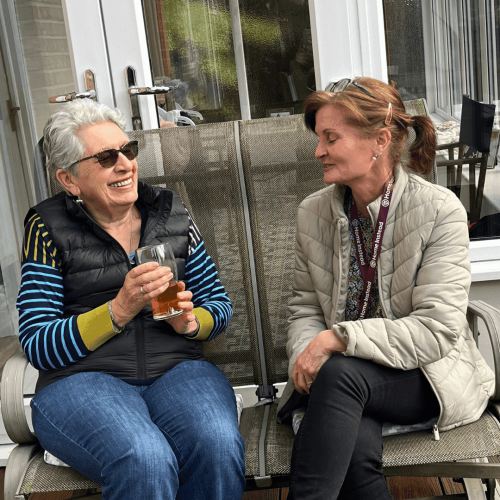 Two women sitting and smiling on a patio, one holding a drink, both wearing jackets, with a glass door behind them. - Home Instead