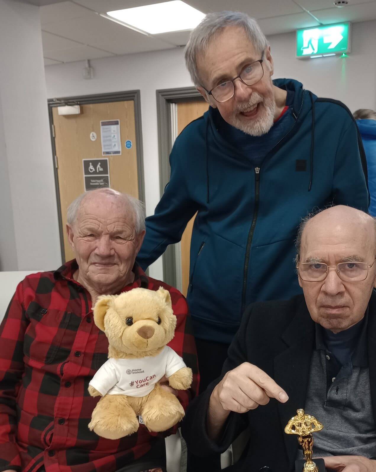 Three elderly men smiling, one holding a teddy bear wearing a shirt; the other holding a small trophy-like object. - Home Instead