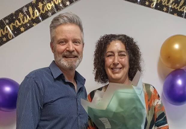 Two people smiling, with one holding a bouquet. "Congratulations" banner and balloons in the background. - Home Instead