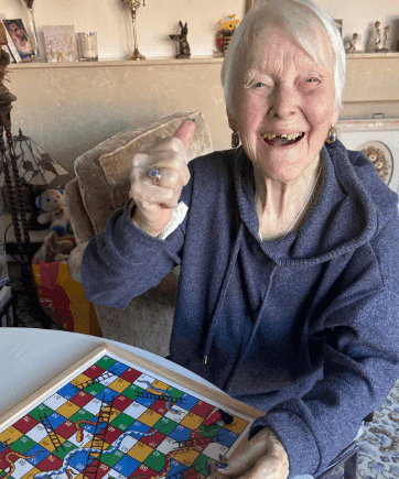 An elderly woman smiling and giving a thumbs up while seated at a table with a colorful board game. - Home Instead