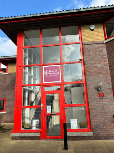 Tall office building exterior with red-framed glass entrance displaying a Home Instead logo and signage on the door. - Home Instead