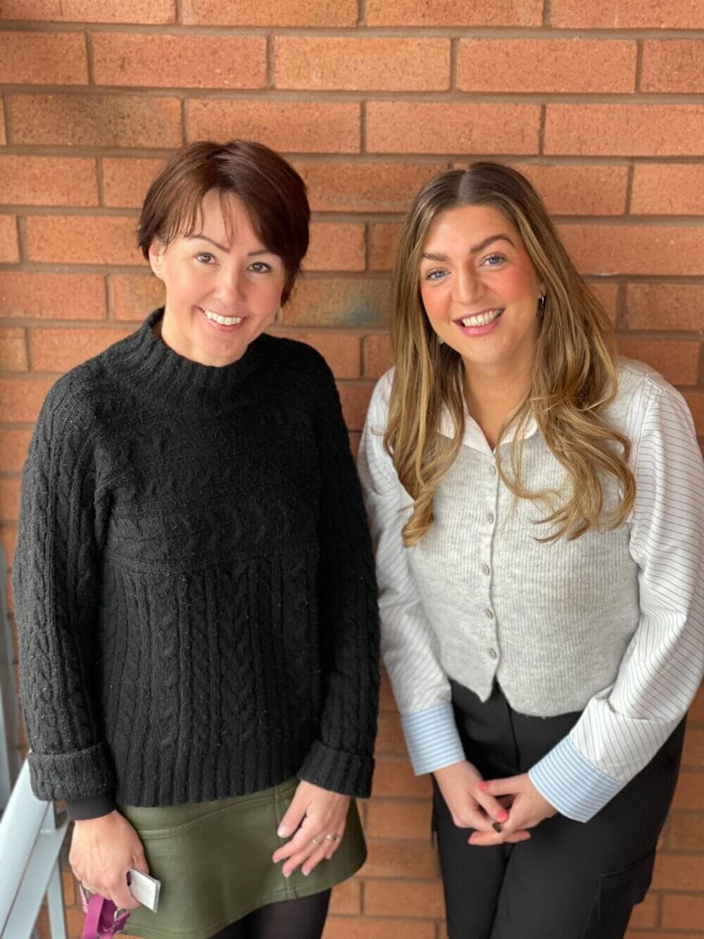 Two women smiling and standing against a brick wall, one in a black sweater and the other in a grey vest. - Home Instead