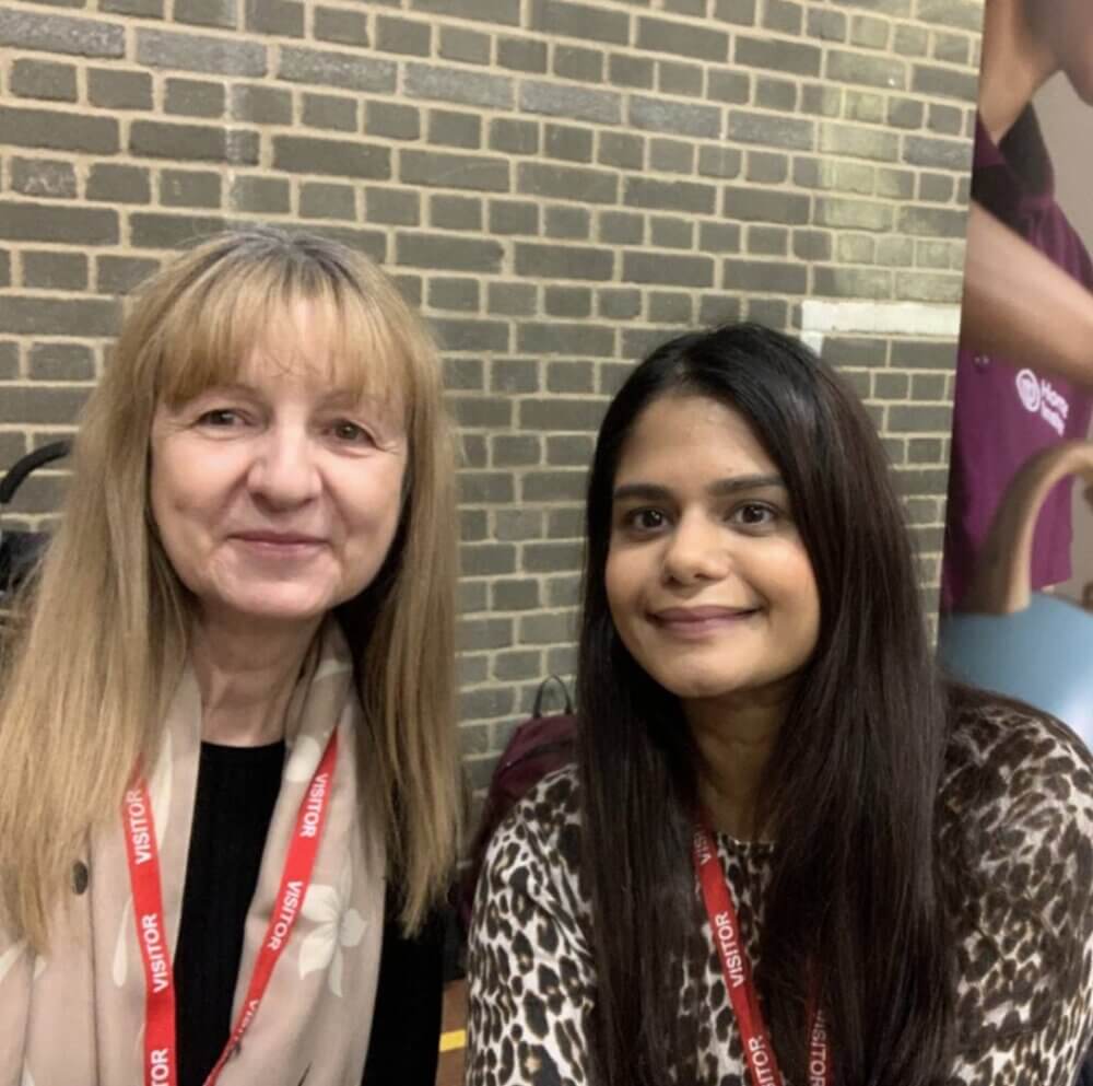 Two women with visitor badges, one with blonde hair and the other with long dark hair, smiling in front of a brick wall. - Home Instead
