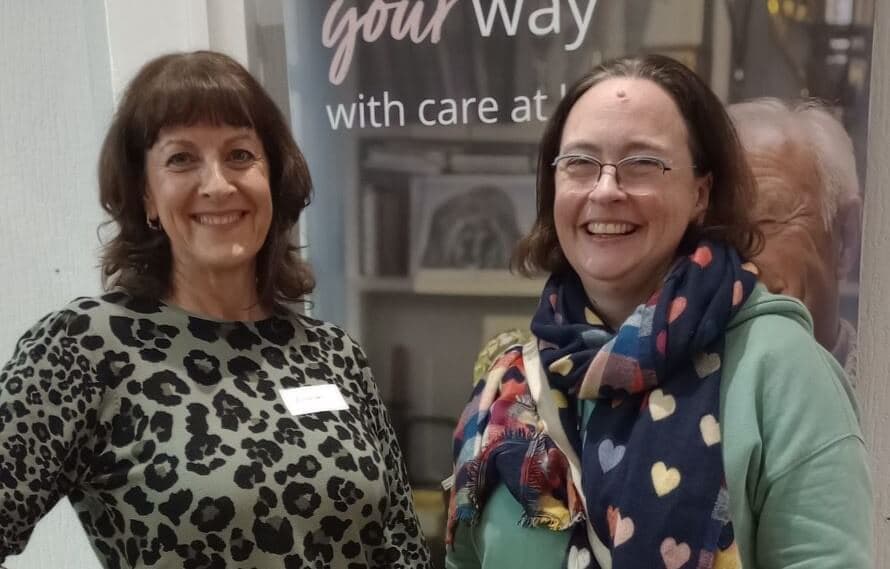 Two women stand smiling in front of a sign. One wears a leopard print shirt, the other a heart-patterned scarf. - Home Instead