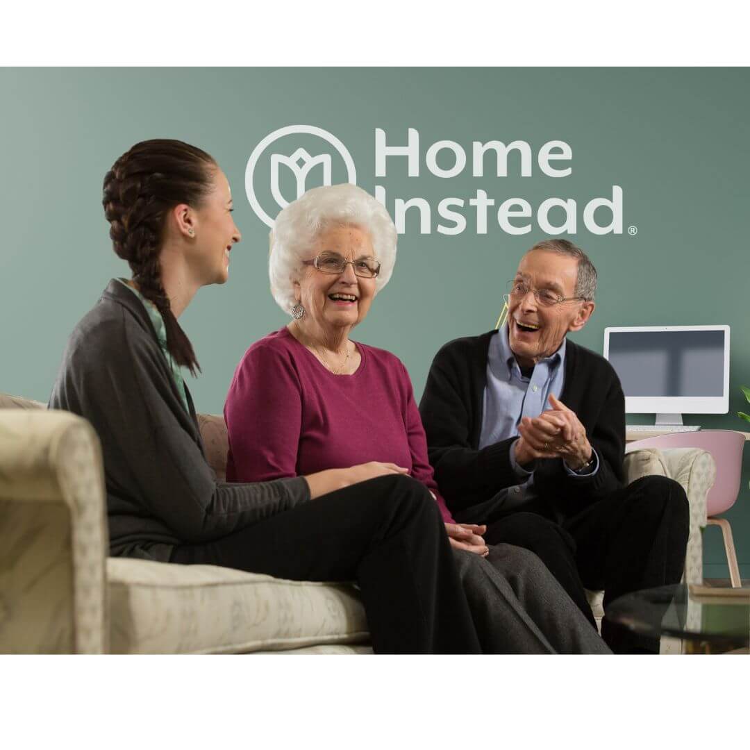 Three people, including an elderly woman, chatting and laughing on a couch with a "Home Instead" logo in the background. - Home Instead