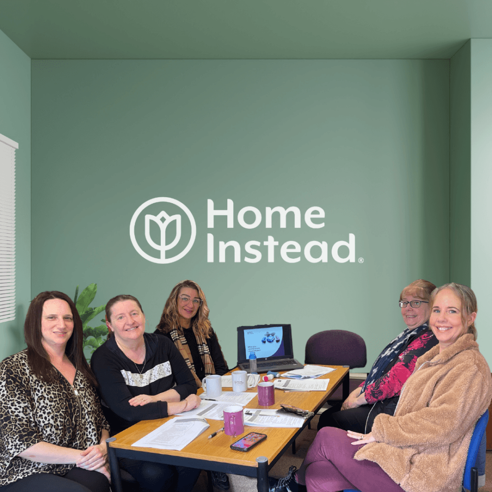 A group of five people sit around a table with a laptop, documents, and coffee mugs. The wall behind them says "Home Instead. - Home Instead
