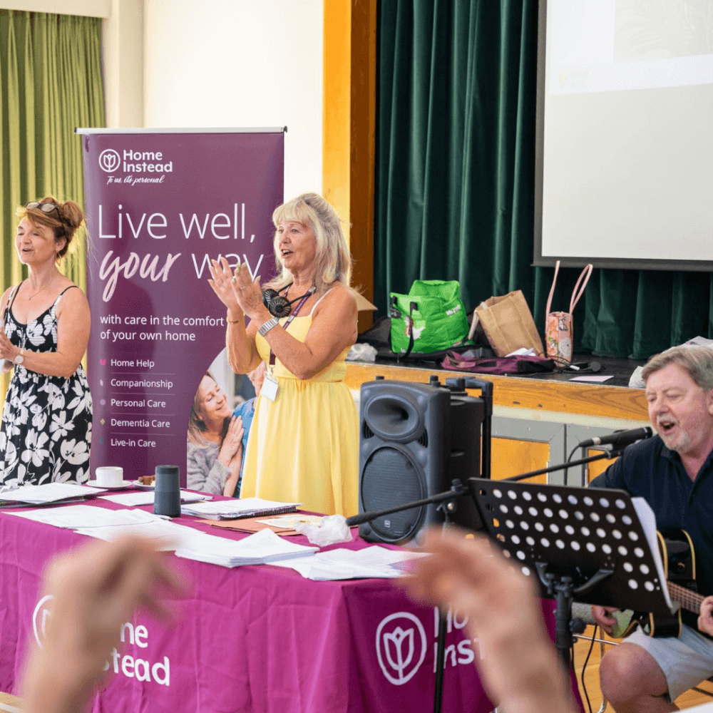 Three people are singing and playing music on a stage with "Home Instead" banner in the background. - Home Instead