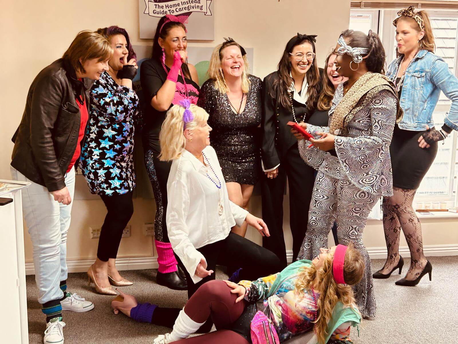 A group of women in colorful '80s outfits laughing and posing together indoors. - Home Instead