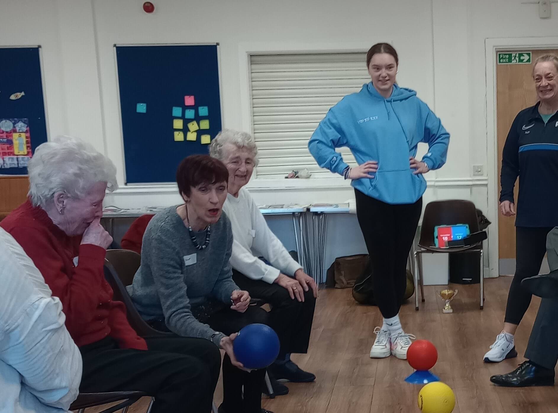 Group of elderly people playing a seated ball game indoors; younger person stands nearby smiling. - Home Instead