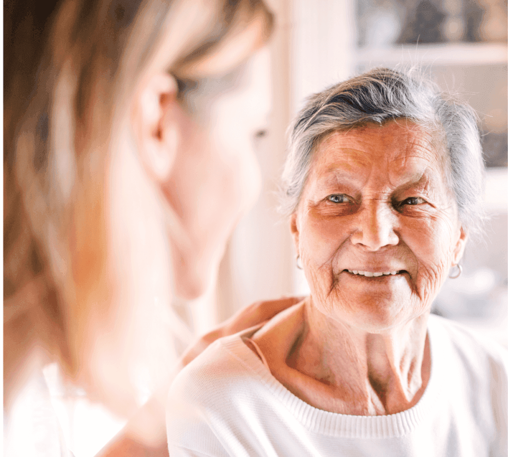 An elderly woman smiles warmly at a younger woman, who has her hand on the elderly woman's shoulder. - Home Instead