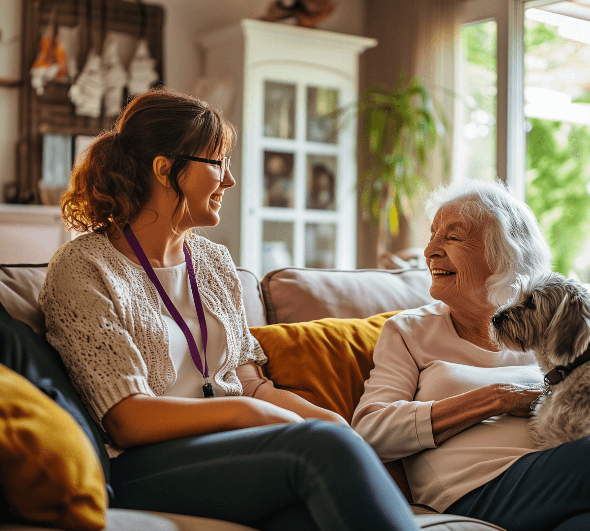 A female Home Instead Care Professional sits on the sofa beside her female home care client who has her dog on her lap. Both are smiling.