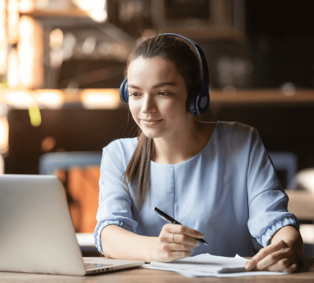 A woman wearing headphones sits at a table, writing notes while looking at her laptop screen. - Home Instead