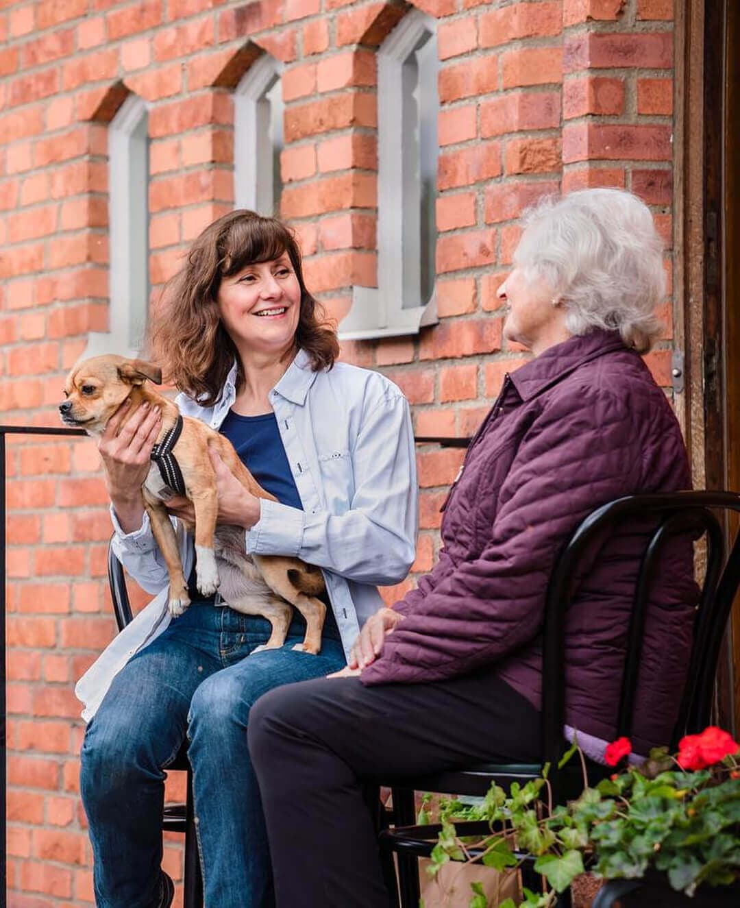 Two women smiling and talking outside with a small dog on one woman's lap, sitting by a brick wall. - Home Instead