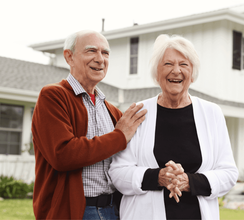 An older male and female couple stand on the lawn outside their house. Both are smiling and the gentleman has his hand on the woman's shoulder.