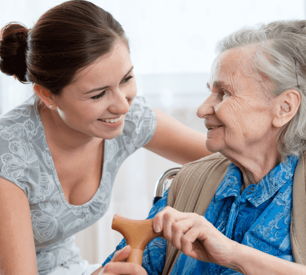 Young woman smiles and talks to an elderly woman with a cane, both showing warmth and connection. - Home Instead
