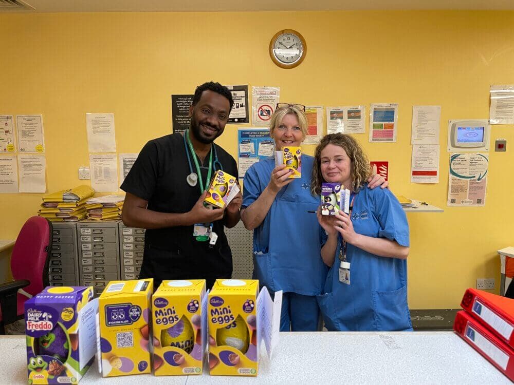 Three smiling healthcare workers holding Easter treats in a hospital break room with more treats on the counter. - Home Instead
