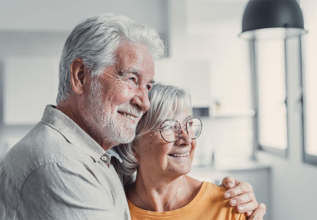 Elderly couple smiling and hugging, standing indoors near a window, with natural light illuminating them. - Home Instead