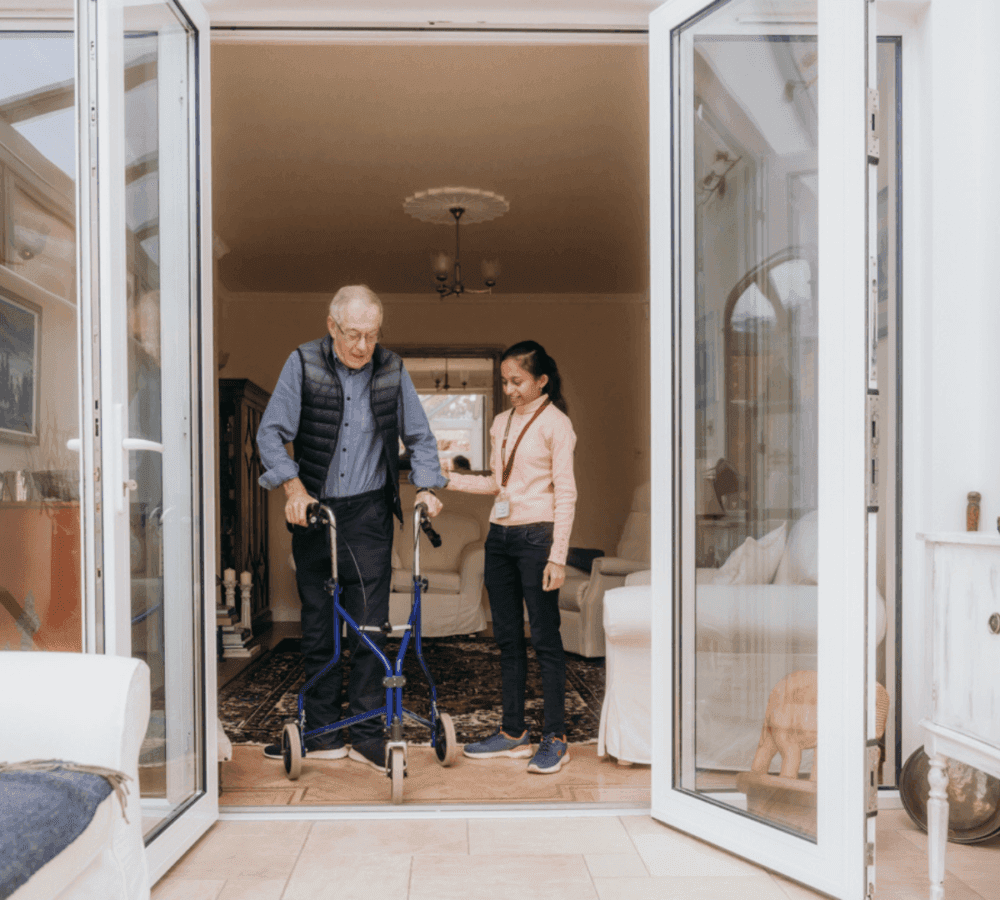 An elderly man using a walker is assisted by a young woman as they walk through a large open doorway. - Home Instead Poole