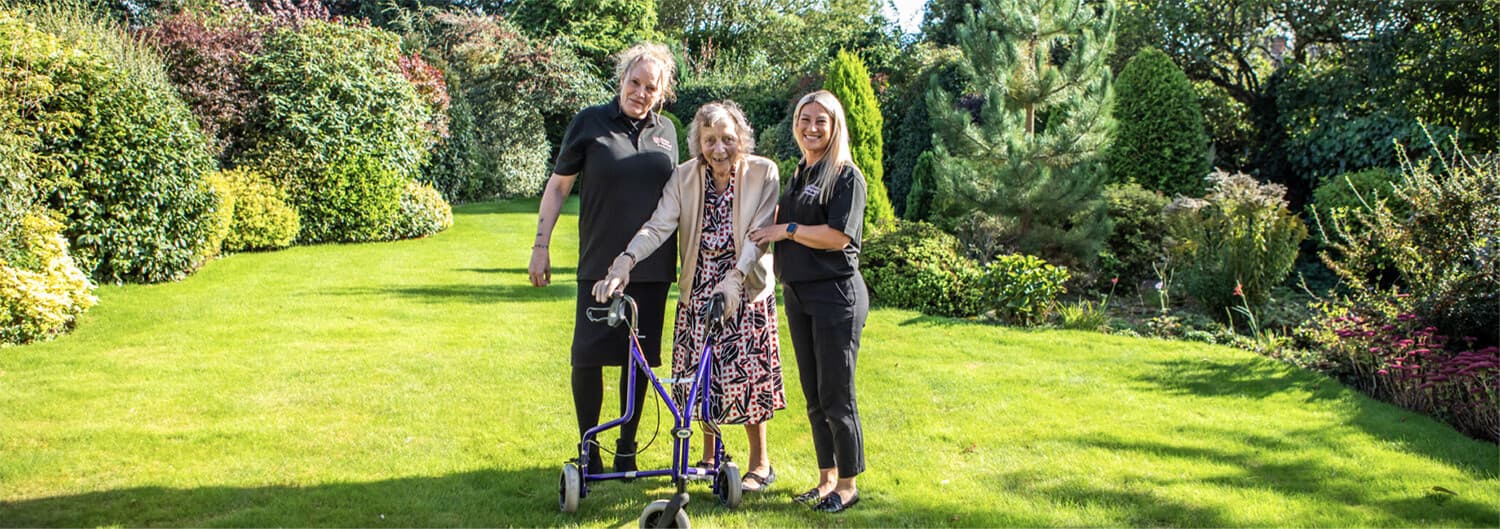 Three smiling people stand in a lush garden, one of them using a walker. - Home Instead