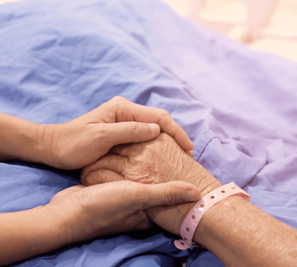A younger hand gently holding an older hand, both resting on a blue blanket, with a pink hospital wristband visible. - Home Instead