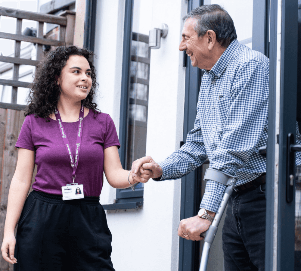 A caregiver shakes hands with a man using crutches outside a building. Both are smiling warmly at each other. - Home Instead