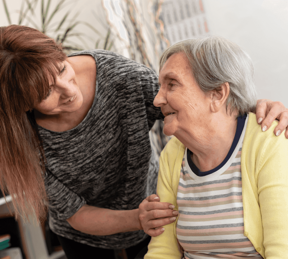 A younger woman affectionately leans over an elderly woman, both smiling and holding each other's shoulders. - Home Instead Poole