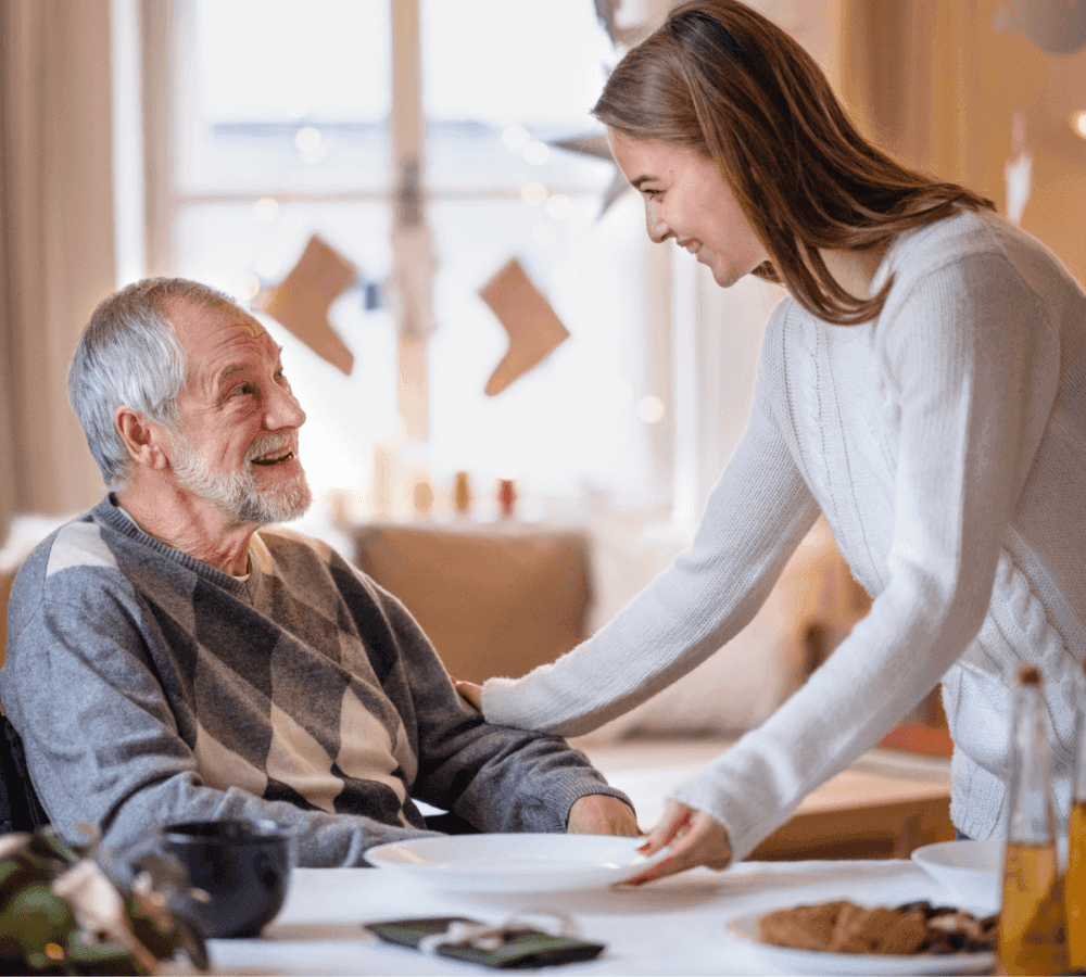 Young woman smiling and serving food to an elderly man at a dining table, both enjoying a cosy, festive setting. - Home Instead Poole