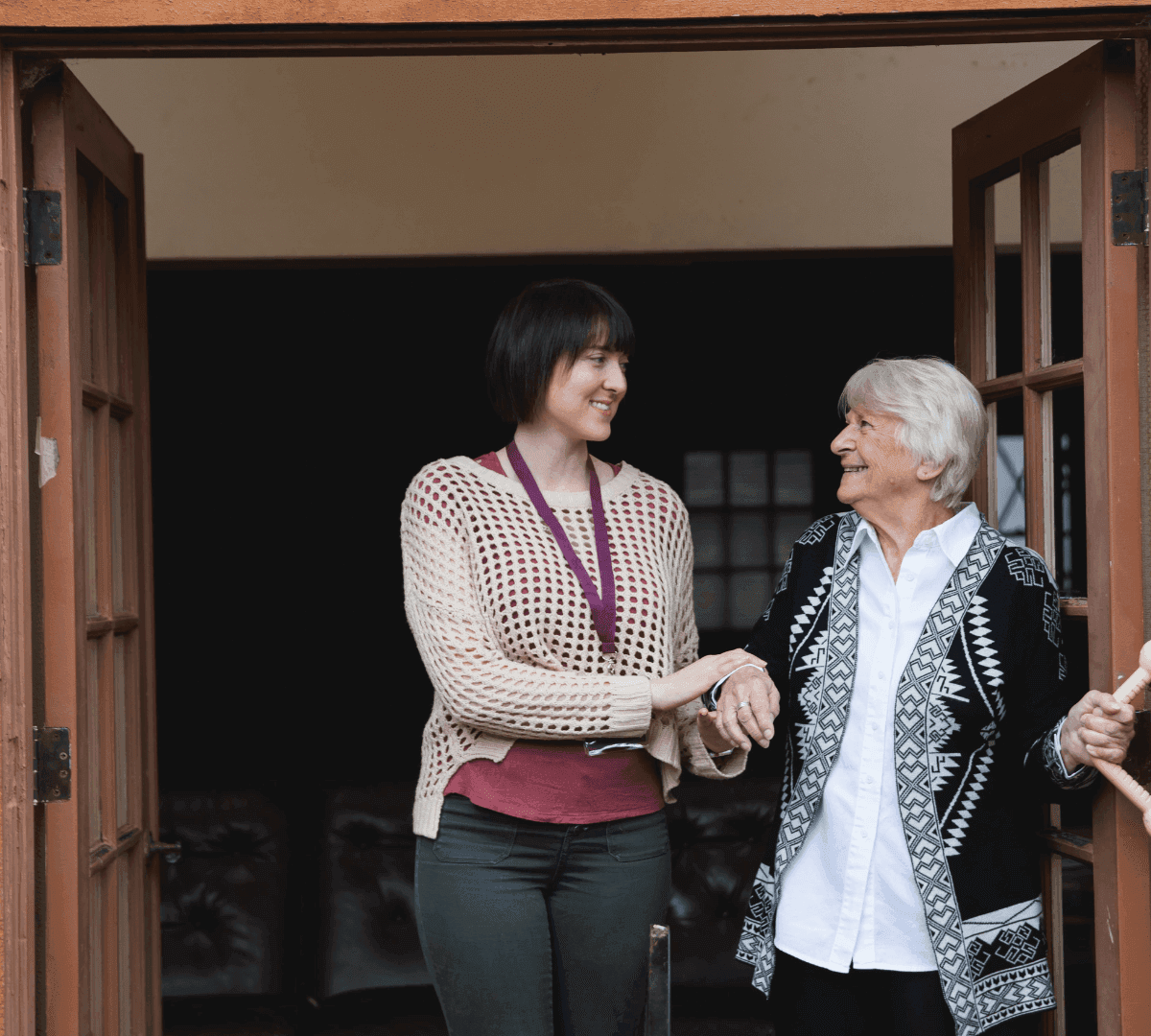 Two women standing in a doorway, one elderly and one younger, holding hands and smiling at each other. - Home Instead