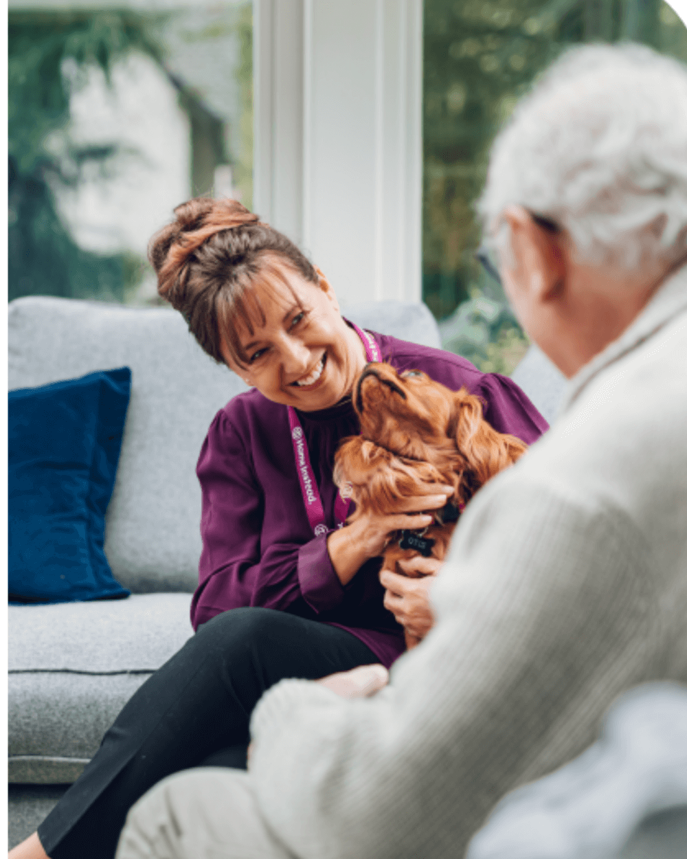 Caretaker in purple shirt and elderly man with white hair smiling at a dog in a cozy, bright room. - Home Instead