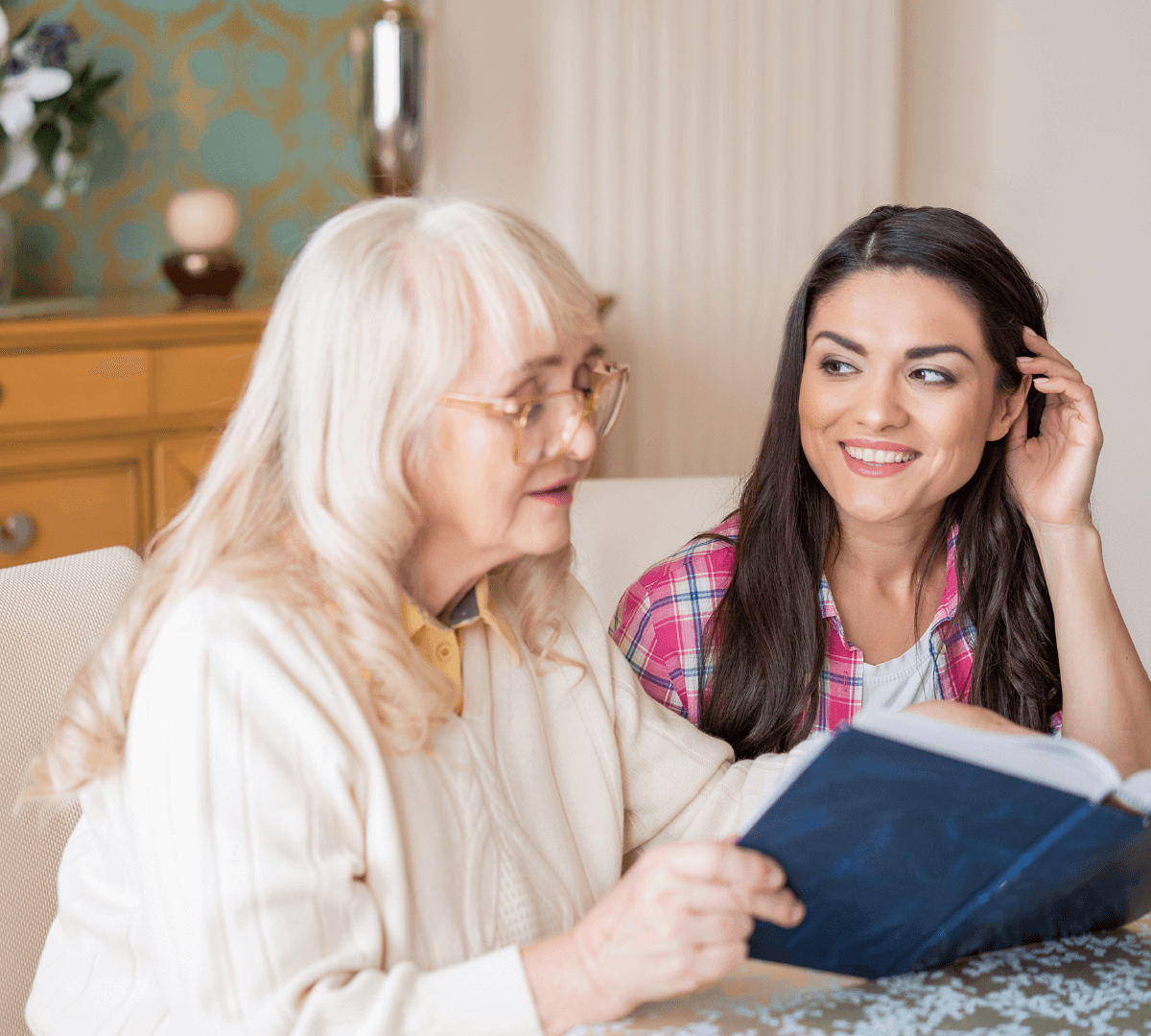 Live in Care in Chesterfield Older lady with long grey blonde hair sitting at home on sofa with a younger Care Professional turning to face her as they share a book