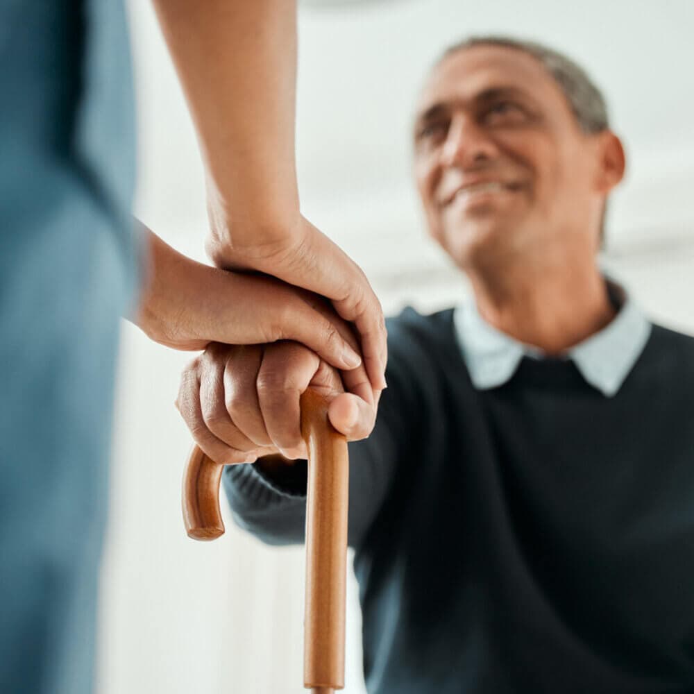 Care professional supports an elderly person by holding their hand on a walking stick