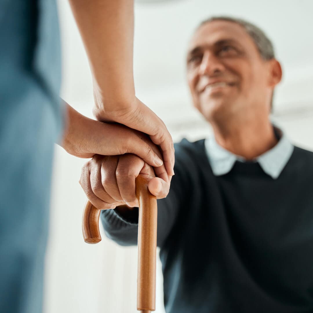 Care professional supports an elderly person by holding their hand on a walking stick