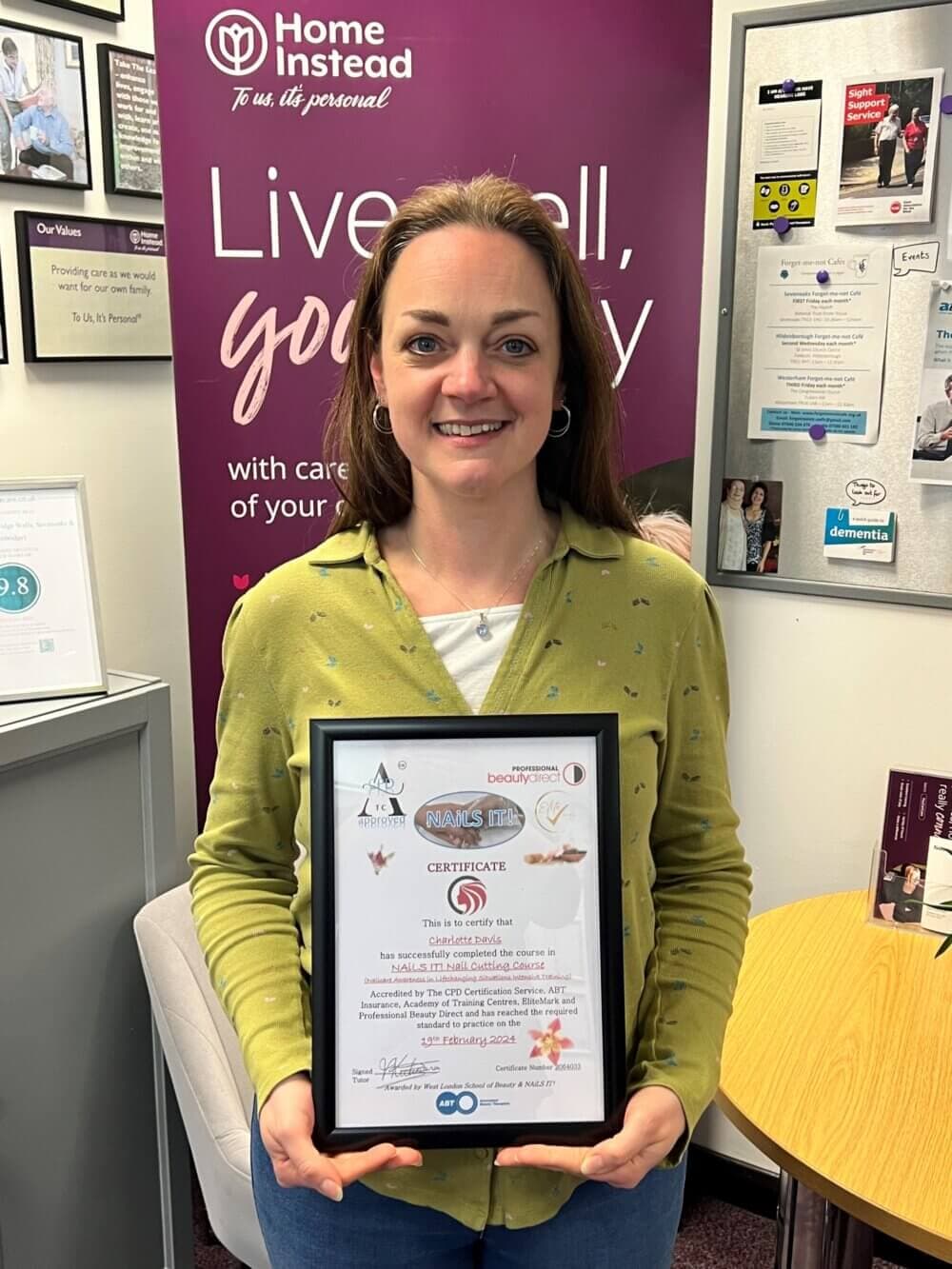 A person smiling while holding a framed certificate in an office with a board behind them displaying various notices. - Home Instead