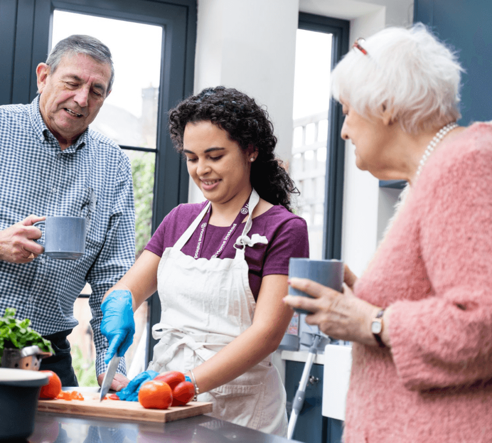 A young woman chops vegetables in a kitchen, while two seniors stand nearby with cups, chatting and smiling. - Home Instead