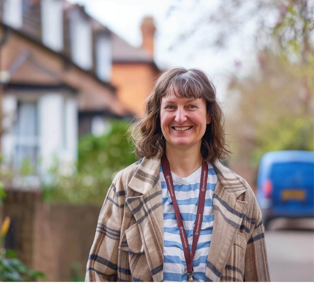 A female Home Instead Care Professional stands in a residential street, smiling at the camera.