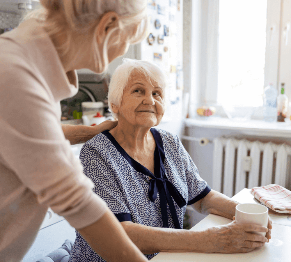Elderly woman sitting at a table with a mug, looking at a standing person touching her shoulder in a sunlit kitchen. - Home Instead Poole