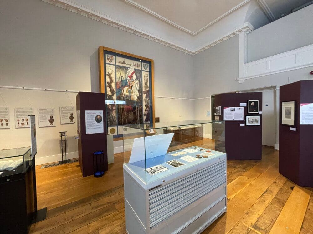 A museum exhibit displaying historical artifacts in glass cases with an illustrated painting on the wall in the background. - Home Instead