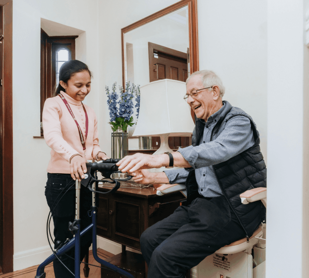 A senior man uses a walker while a smiling caregiver assists him inside a cosy, well-lit room. - Home Instead Poole