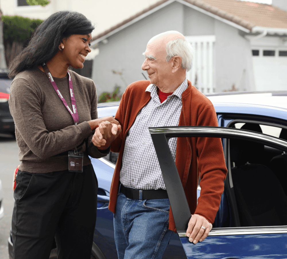 A smiling woman helps an elderly man standing beside a car, holding his hand supportively in a friendly neighbourhood. - Home Instead Southampton