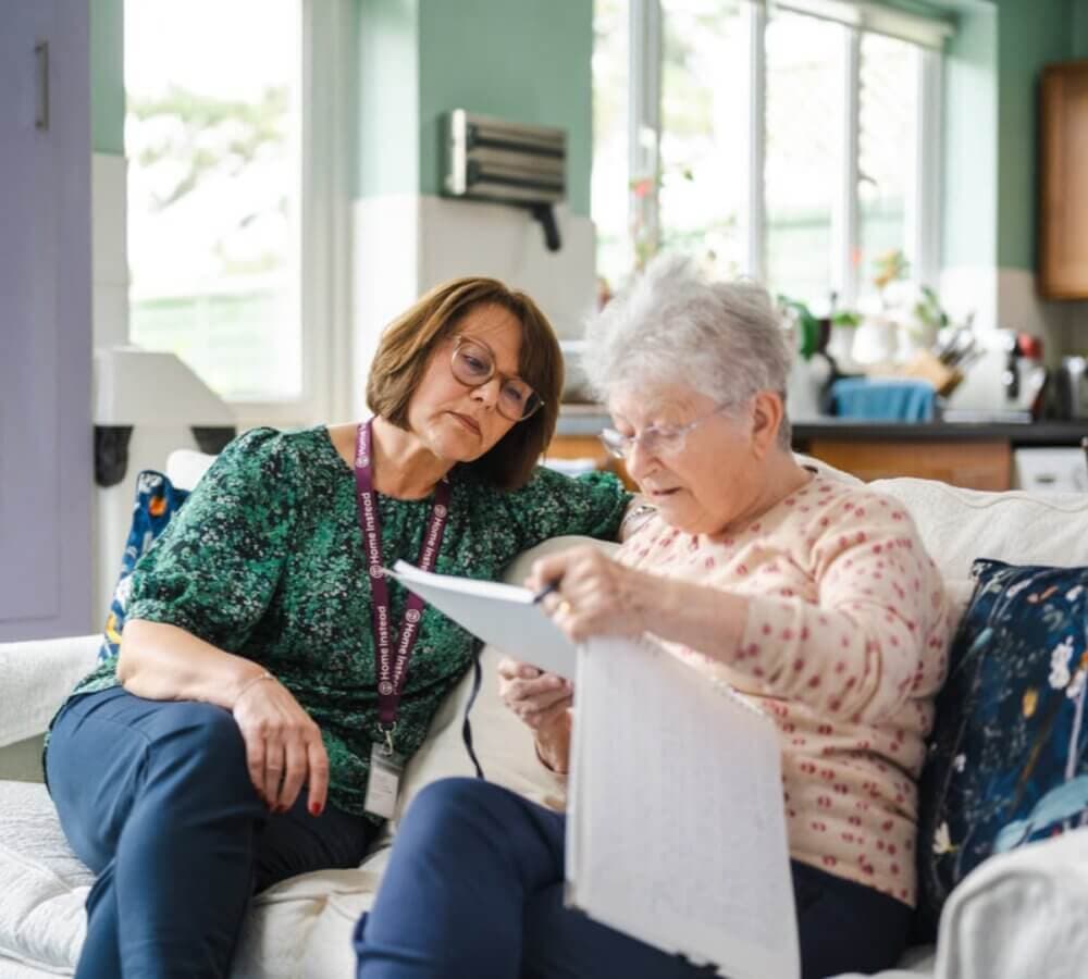 A female Home Instead Care Professional sits on the sofa beside her older female home care client as they read through a booklet the client is holding.