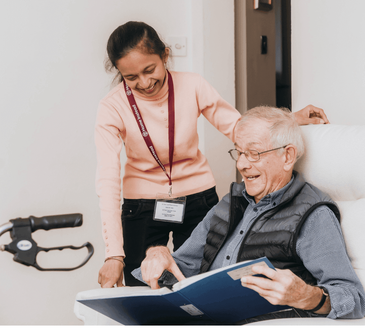 A caregiver smiling and assisting an elderly man who is reading a book while seated in a chair. - Home Instead Poole