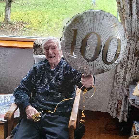 An elderly man smiling and holding a helium balloon with "100" printed on it, sitting indoors near a window. - Home Instead